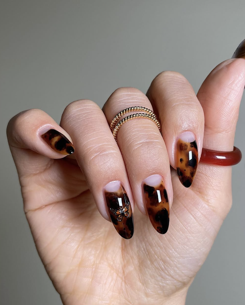 Long, almond-shaped nails featuring a rich, glossy tortoiseshell pattern with an intricate orange and black monarch butterfly charm accent on the ring finger.