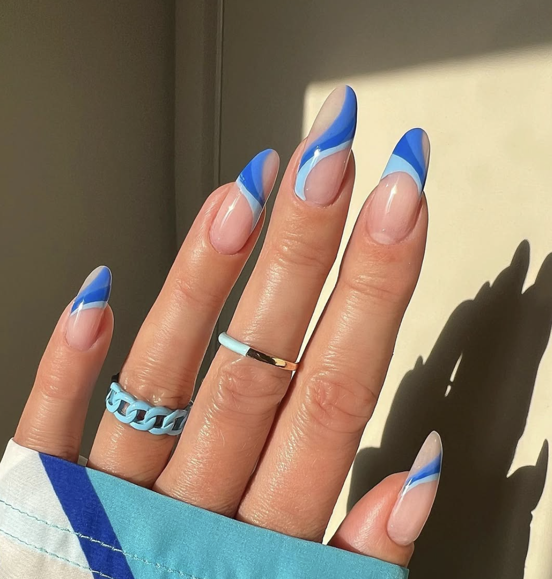 A close-up of almond-shaped nails featuring a minimal nude base with abstract, dual-toned royal blue and sky blue swirl patterns, accessorized with mismatched blue and gold rings.