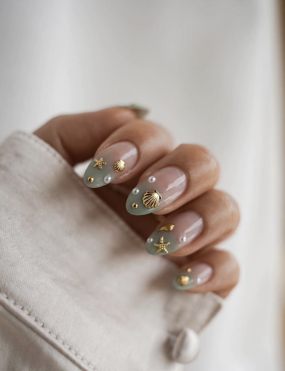 A close-up of long, oval-shaped nails with a sheer nude base, decorated with a sea-glass green French tip outline, delicate 3D gold stars, small gold starfish, and tiny pearl accents, paired with a white linen sleeve.