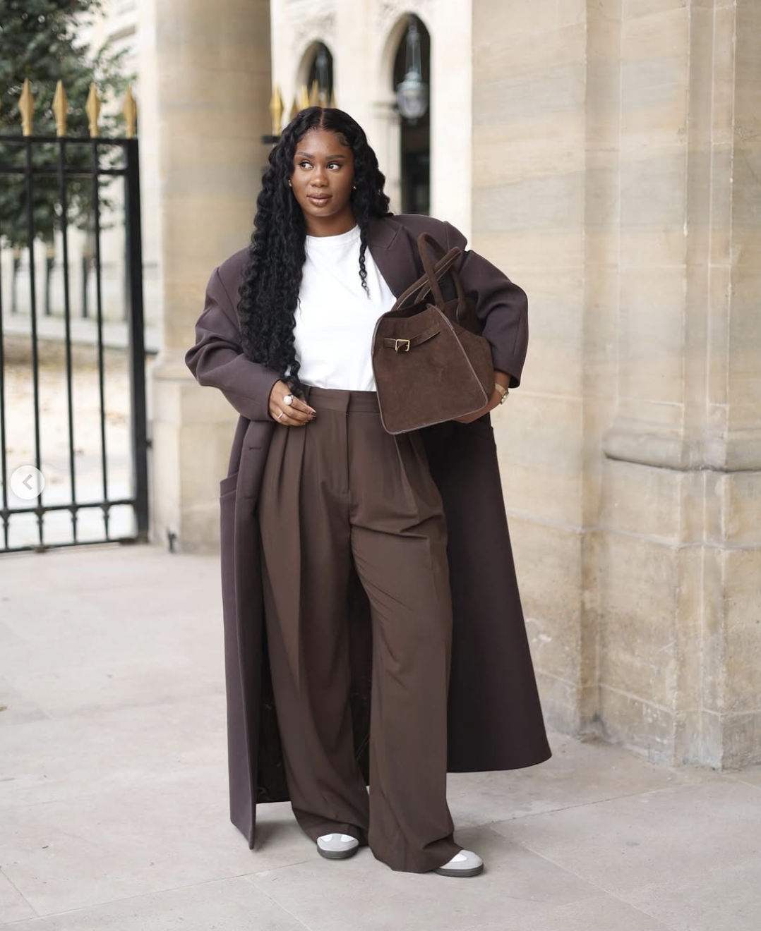 A woman wearing a monochromatic chocolate brown outfit featuring high-waisted wide-leg trousers, a matching floor-length wool coat, and a brown suede handbag.