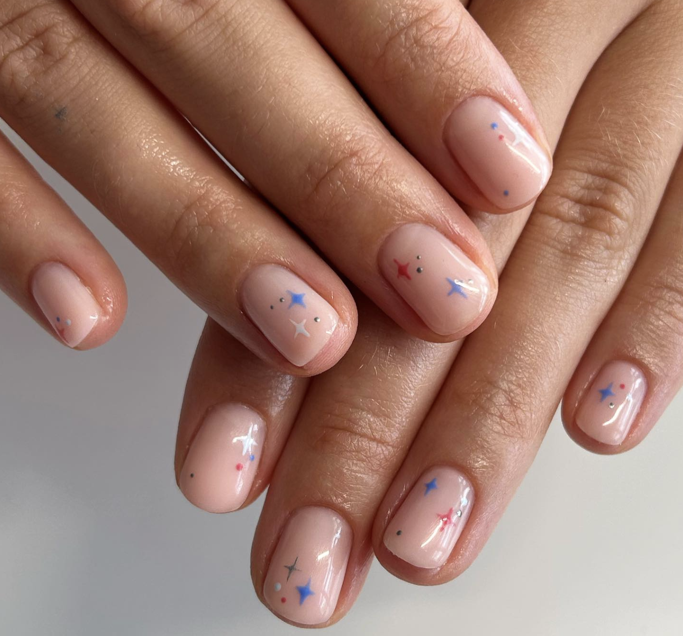 Short natural nails with a sheer pale pink base featuring tiny, colorful retro starburst and micro-dot accents in shades of blue, red, and silver.