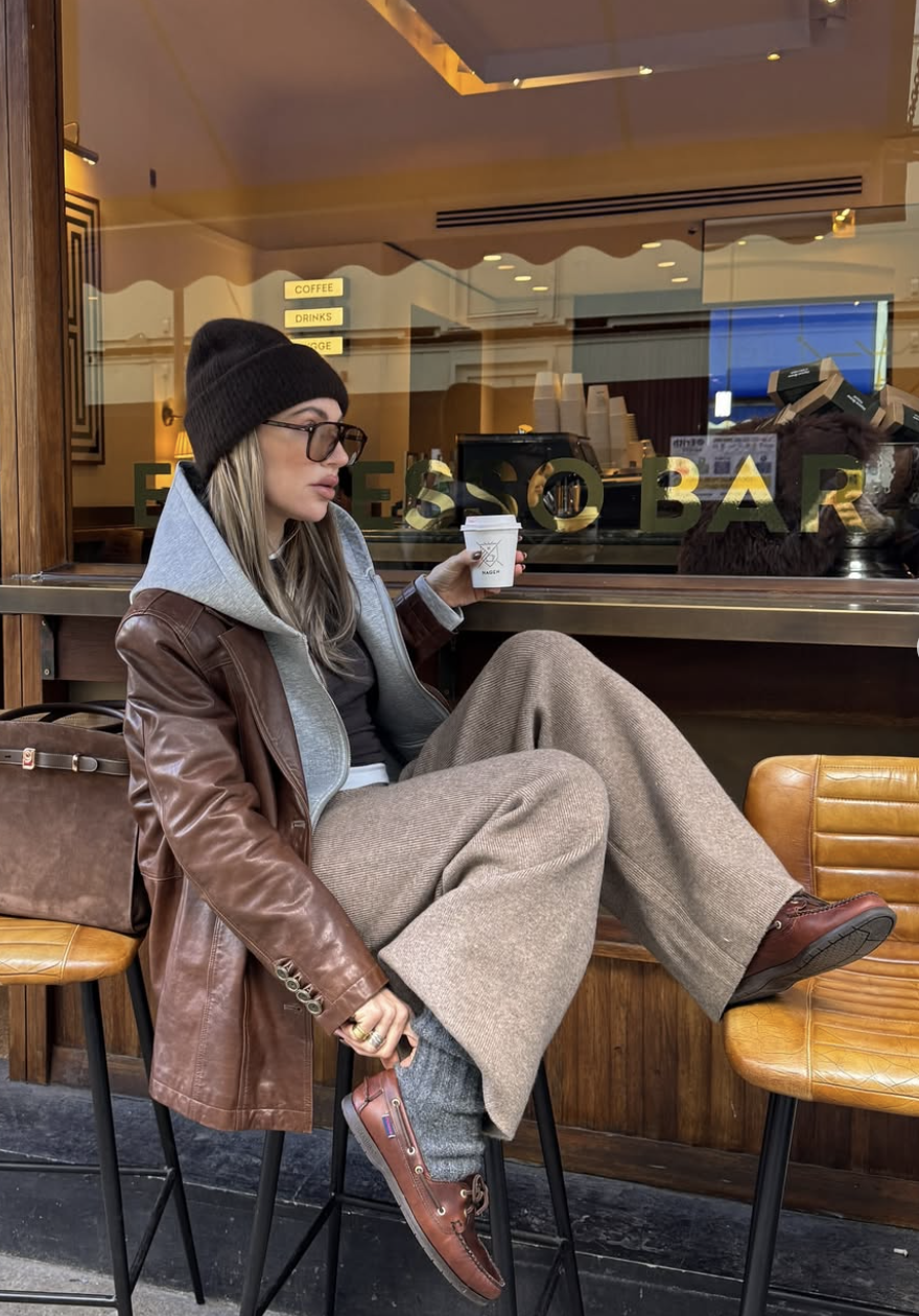 A woman wearing a brown leather blazer layered over a grey hoodie and tan wide-leg trousers, holding a coffee cup for a casual street-style look.