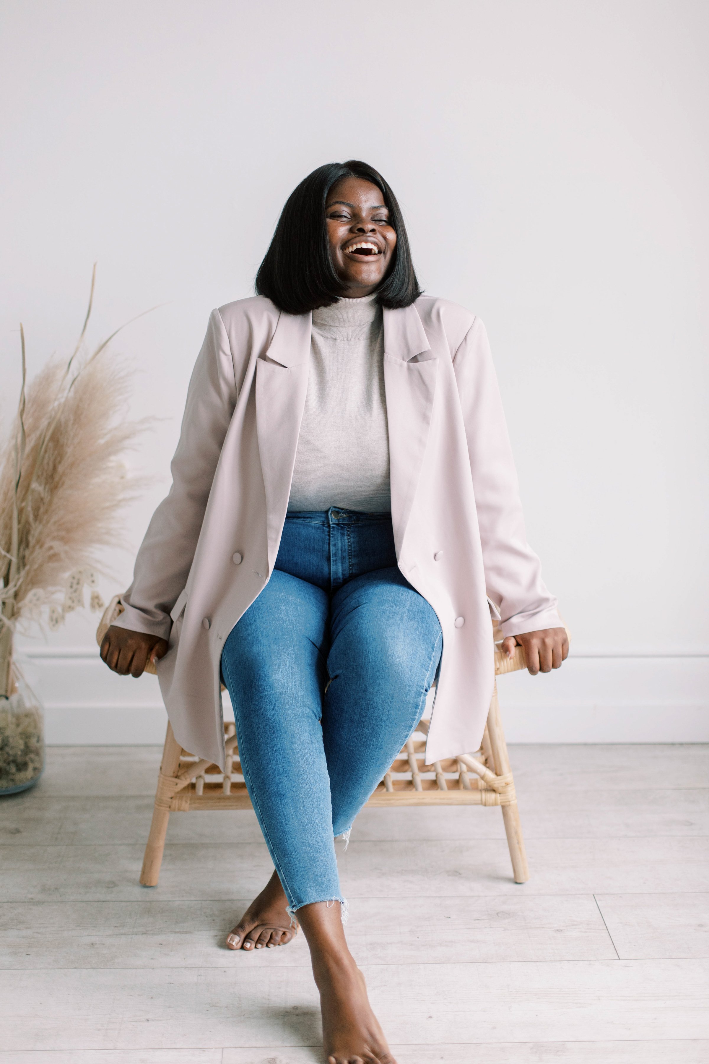 A woman sitting on a wicker chair, laughing, wearing a light-colored blazer, gray turtleneck, and blue jeans, with a white wall and dried plants in the background.