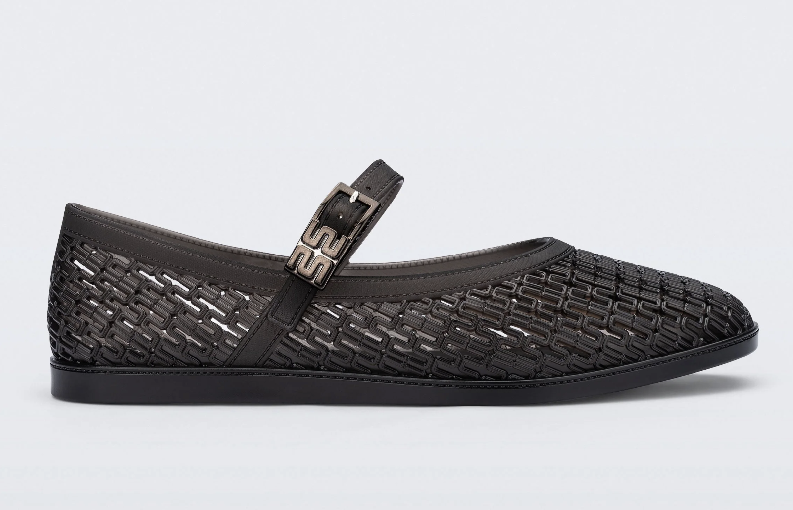 A product view of a dark brown or charcoal grey woven mesh-style jelly Mary Jane flat shoe featuring a rounded toe, a flexible lattice pattern, and a single foot strap with a square buckle.