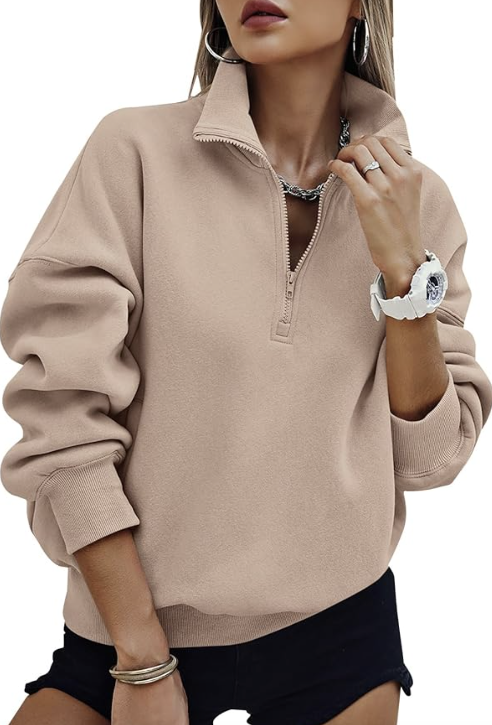 A woman wearing a tan oversized half-zip sweatshirt over a white t-shirt, accessorized with silver bangles and styled with black shorts.