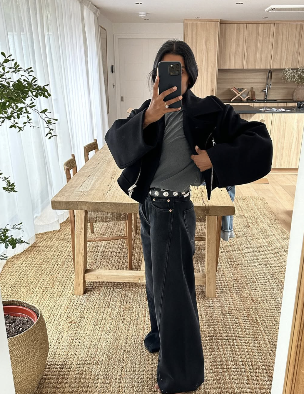 A woman wearing high-waisted black wide-leg jeans with a eyelet grommet belt, a gray top, black outerwear, and pointed heels inside a modern living room.
