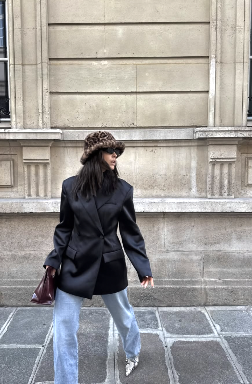 A woman wearing a sleek black bodysuit paired with light grey wide-leg knit pants under a structured black blazer, carrying a mahogany leather tote bag.