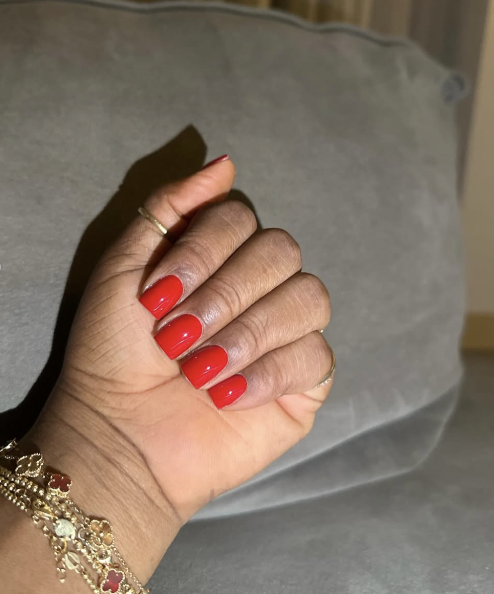 Close-up of short, squoval nails painted in a glossy oxblood red polish with gold bracelets and thin rings.