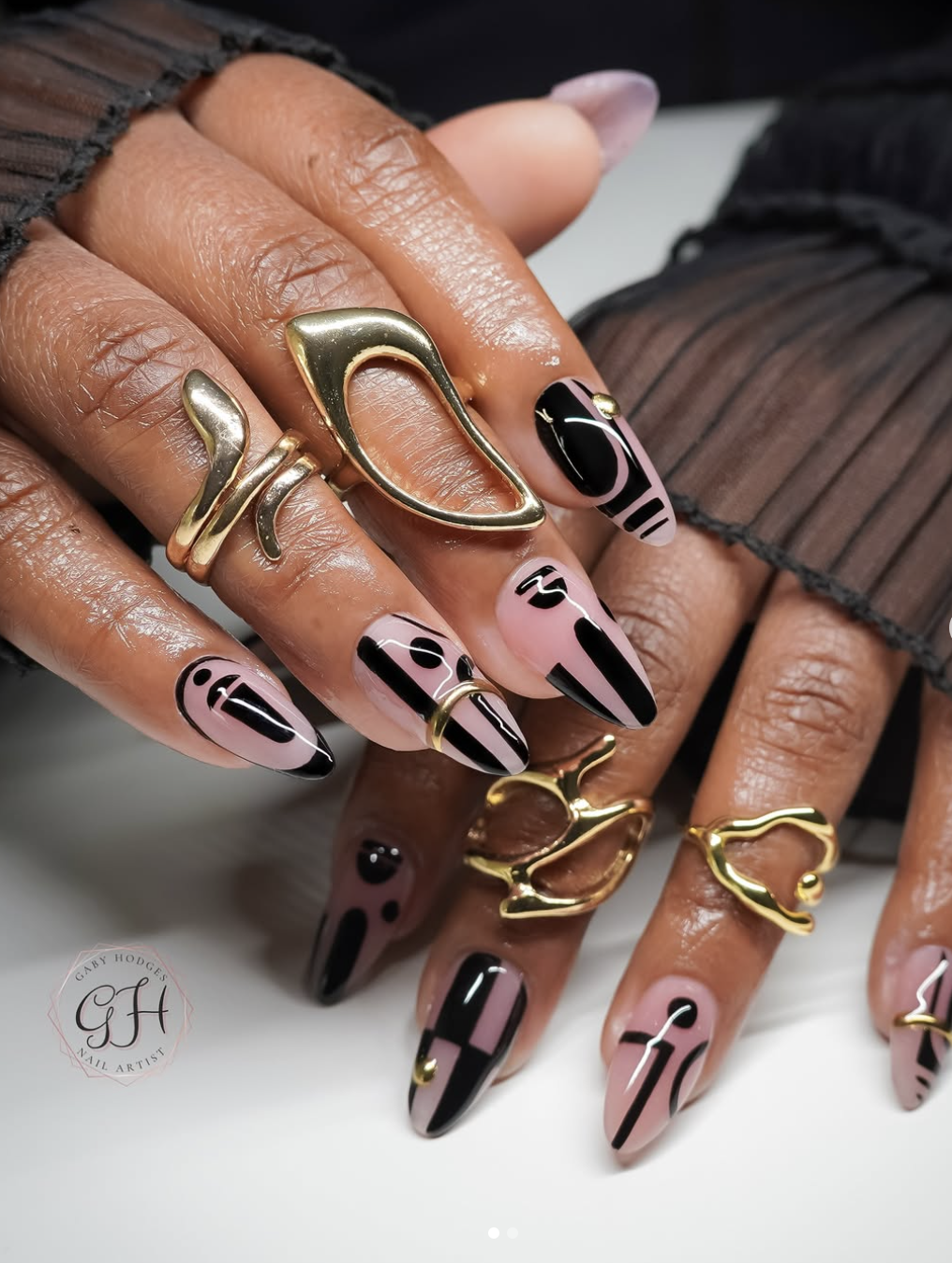 Close-up of a hand with long, almond-shaped pink nails featuring a bold abstract black swirl design and geometric graphic accents, accessorized with heavy, molten-style gold statement rings.