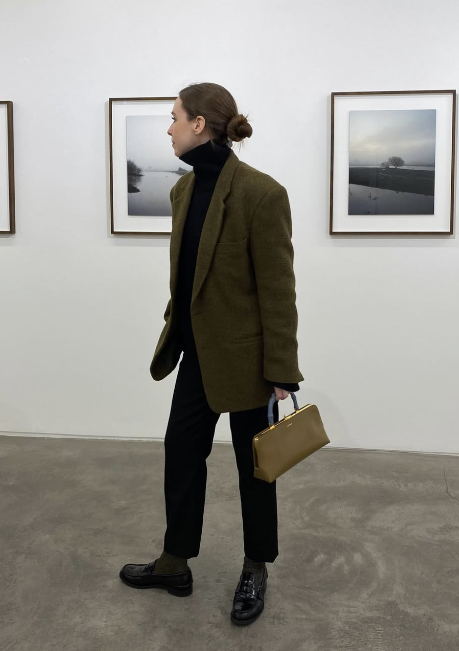 A woman wearing a black turtleneck fitted knit top and dark denim jeans, layered under an olive green structured blazer, accessorized with a tan handbag and black loafers.