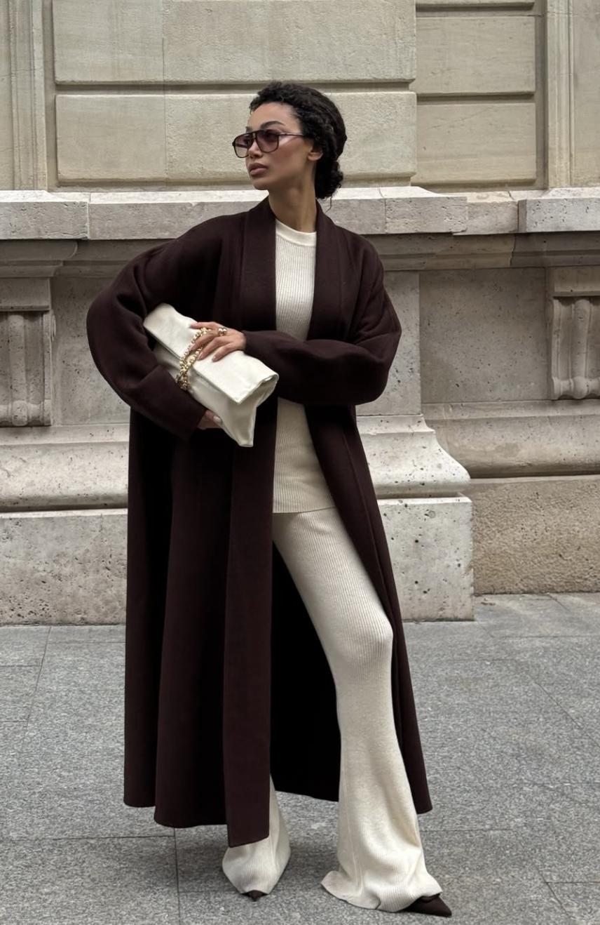 A woman wearing a long chocolate brown coat over an all-white outfit with white straight-leg jeans, accessorized with neutral pointed-toe heels and a white clutch bag.
