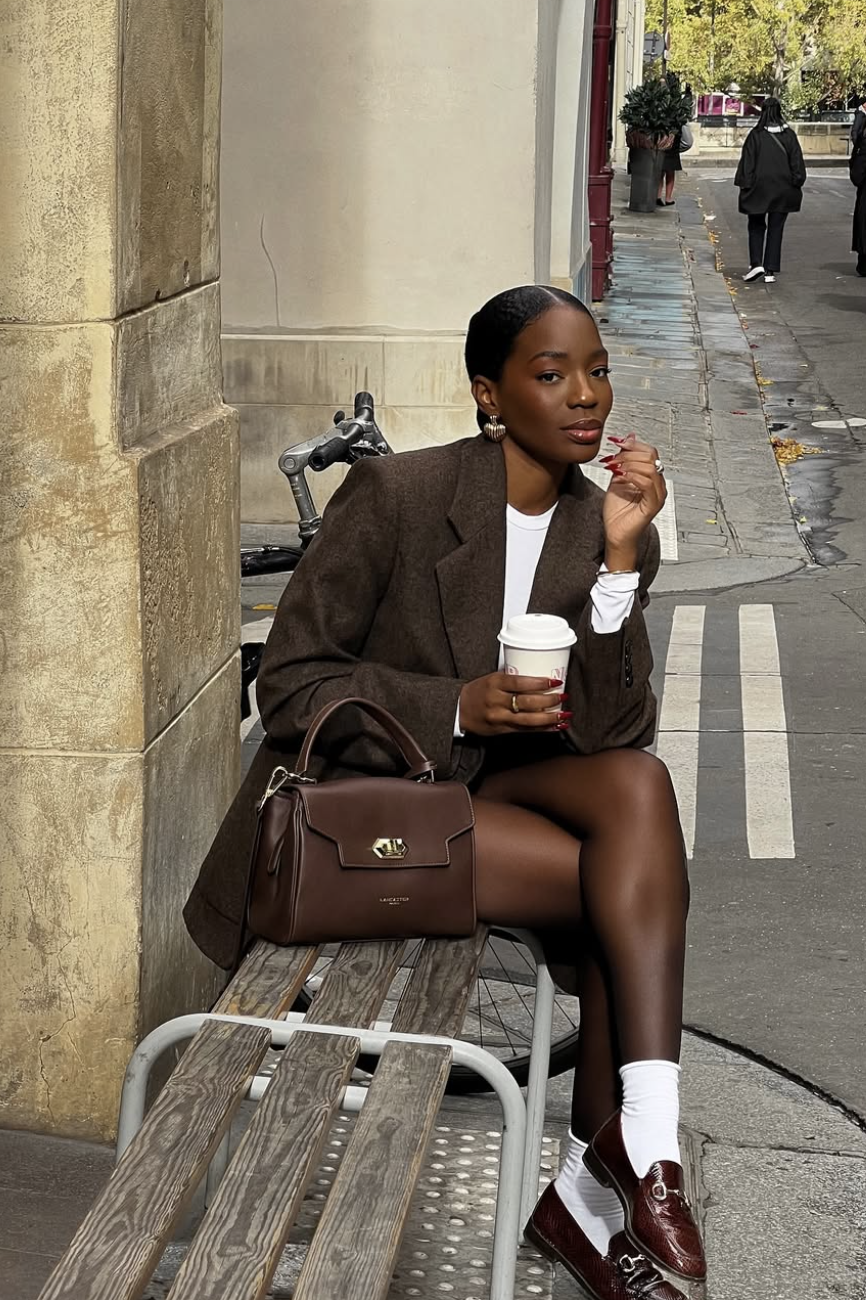 A woman sitting on a wooden bench wearing an oversized dark brown blazer over a white t-shirt, paired with white socks, burgundy loafers, and a structured brown top-handle handbag.