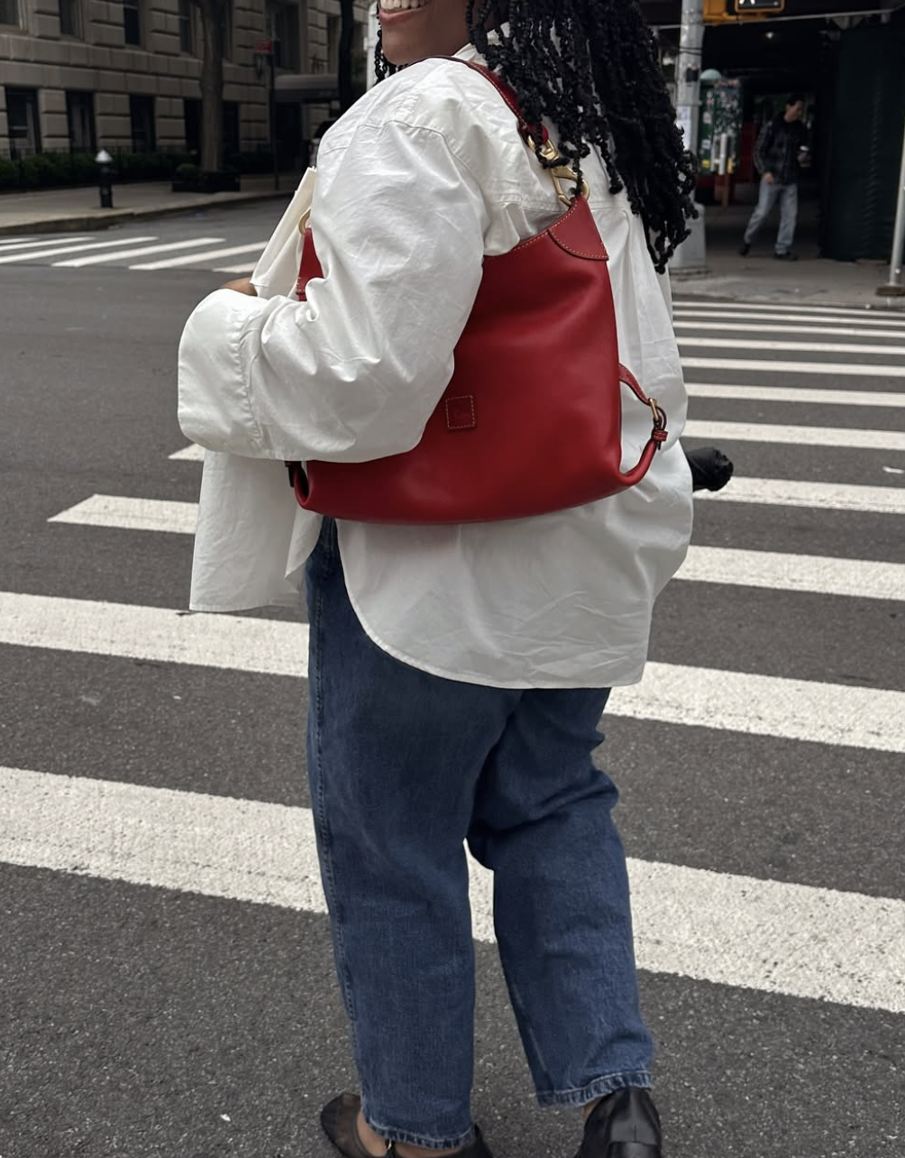 A woman wearing an oversized white button-down shirt, dark wash jeans, and black mesh ballet flats, carrying a large red slouchy shoulder bag.
