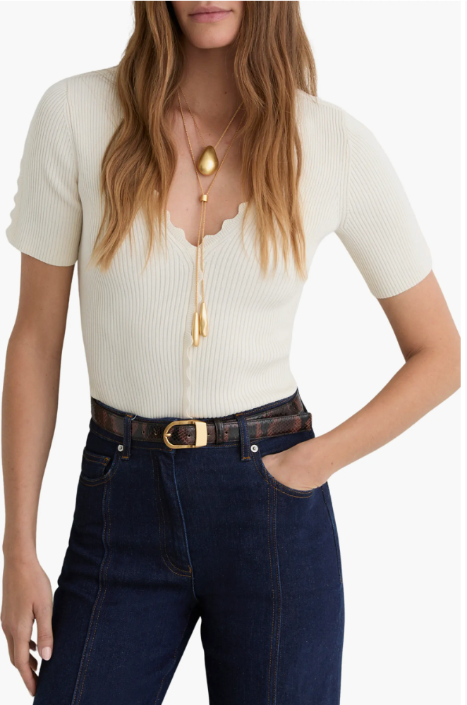 A woman wearing a white short-sleeve rib-knit top with a scalloped V-neckline, styled with dark wash jeans, a black leather belt, and long gold pendant necklaces.