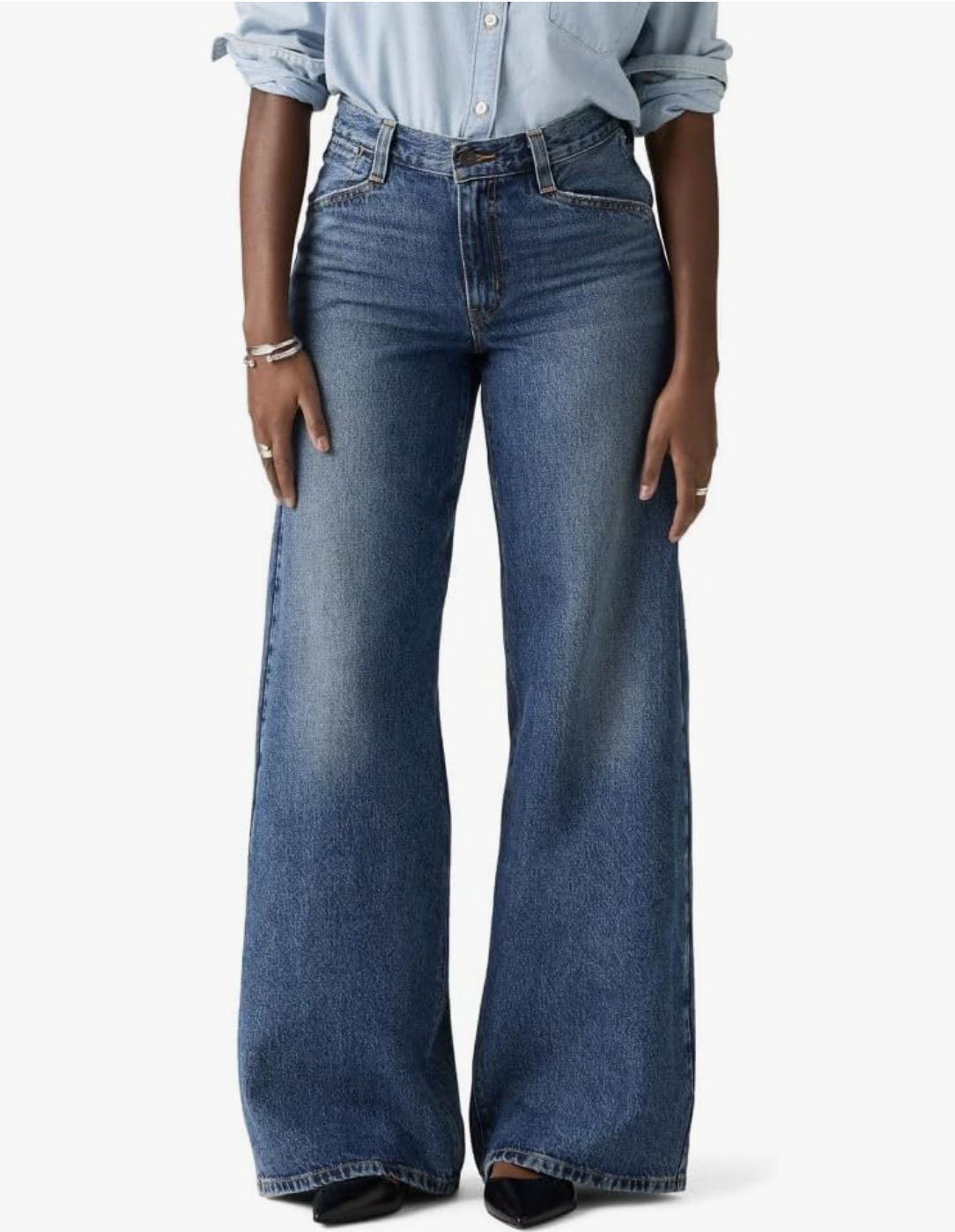 A woman in her 60s wearing Levi's 94 baggy wide-leg jeans in a mid-to-dark wash, styled with a light blue button-down shirt and silver bracelets for a modern spring outfit.