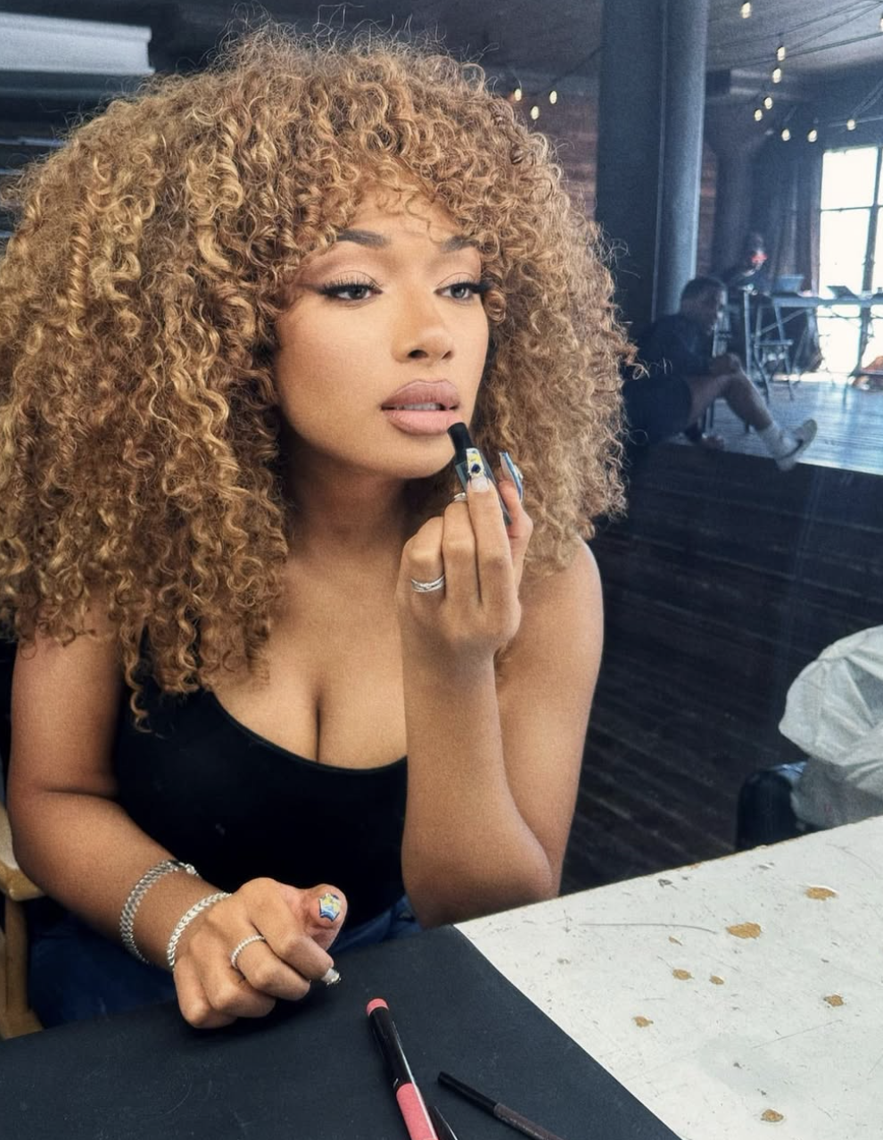 A woman with voluminous, honey-blonde curly hair and dark roots applying lipstick, showcasing a flattering warm hair color for medium and tan skin tones.