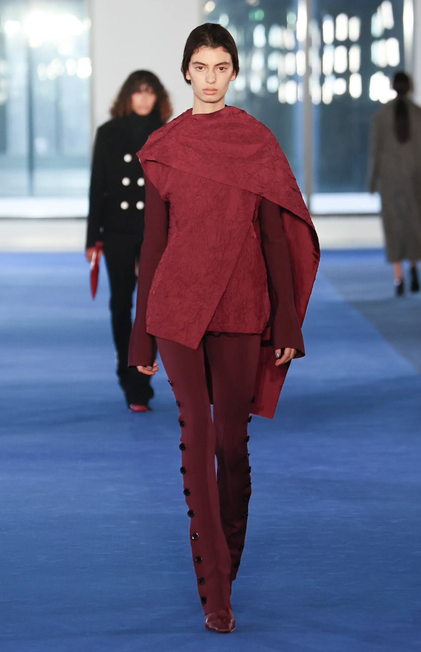 A model on the Proenza Schouler Fall/Winter 2026 runway wearing a structured mahogany red-brown ensemble, illustrating the season's muted clay color trend.
