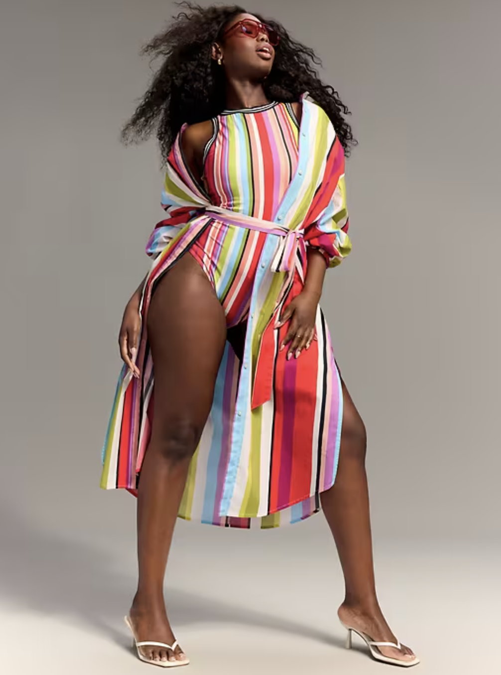 Model wearing the Old Navy x Christopher John Rogers multi-color vertical stripe halter swimsuit and matching oversized striped button-down shirt with white heels.