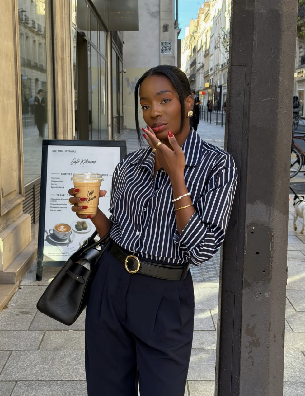 A woman wearing a navy and white striped button-down shirt tucked into navy pleated trousers with a black leather belt.