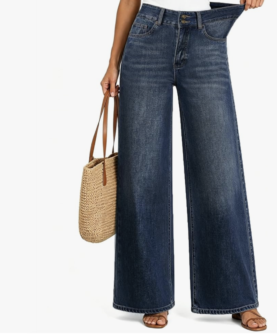 Close-up of a woman wearing high-waisted, dark-wash wide-leg denim jeans with a double-button closure, holding a woven straw tote bag by her side.