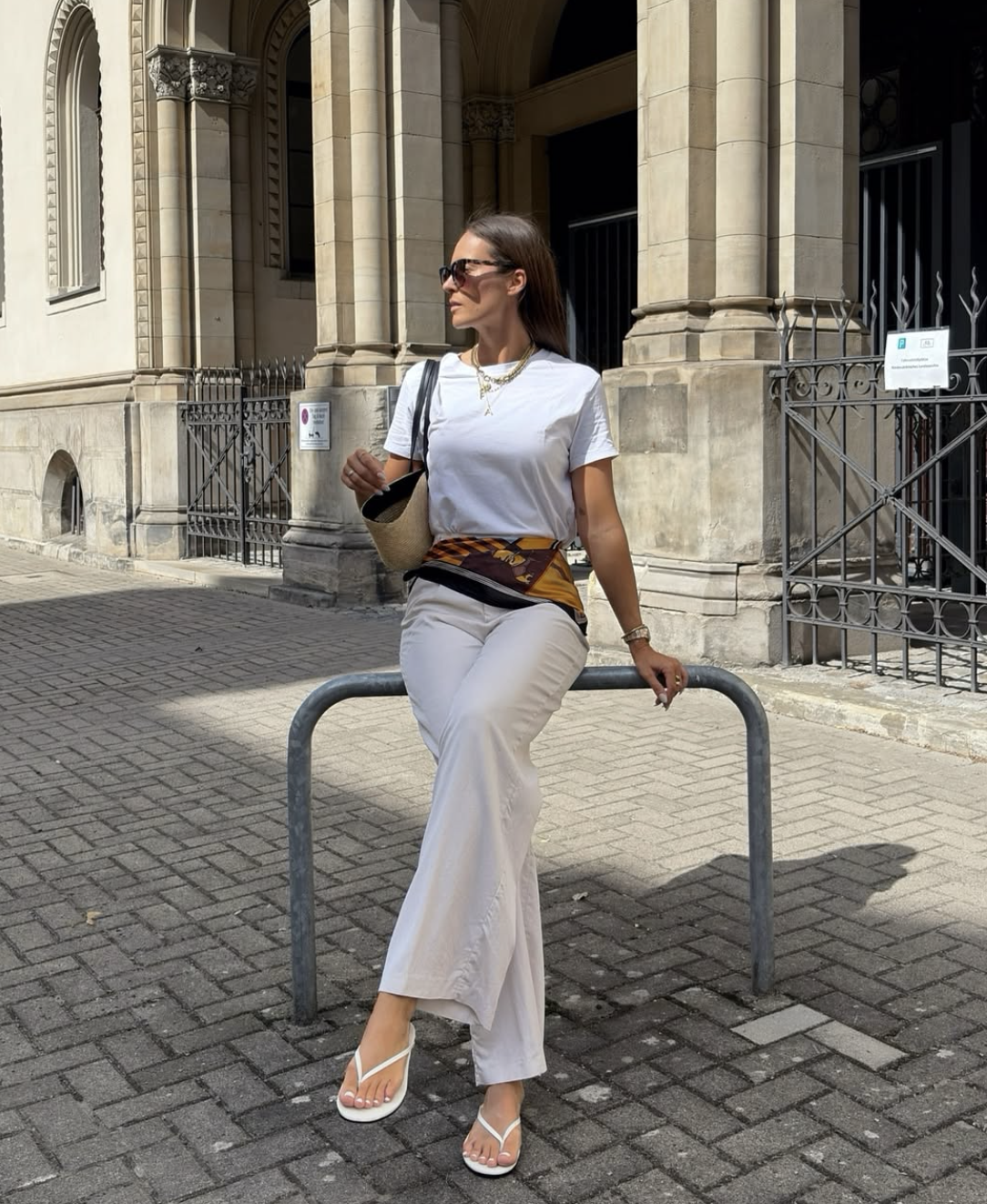 A minimalist all-white summer outfit with a white t-shirt and wide-leg trousers, featuring an amber and chocolate patterned silk scarf tied around the waist.