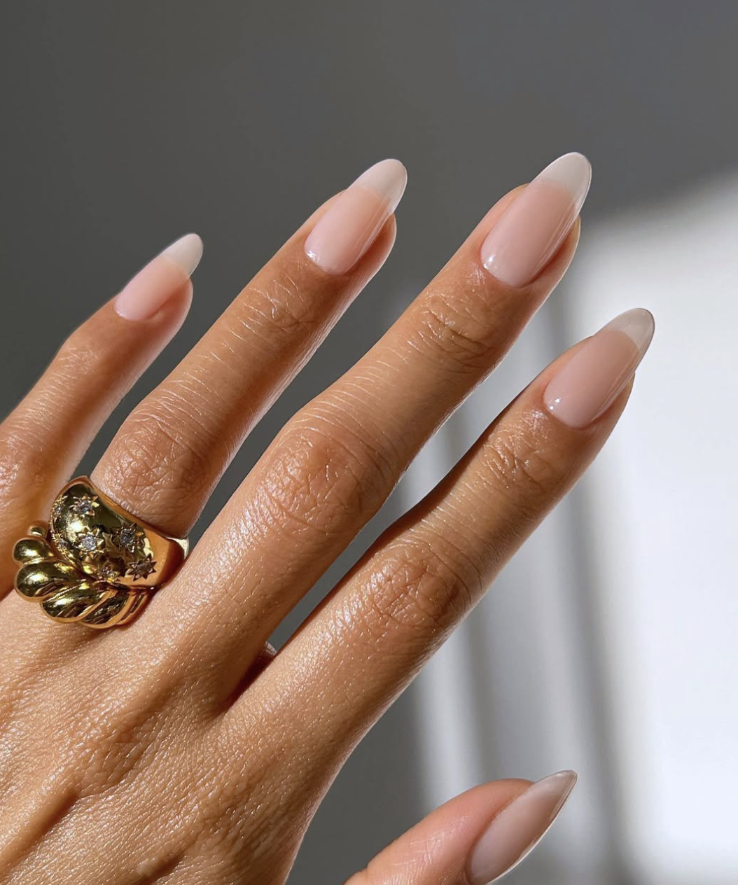 Close-up of a hand with long, almond-shaped nude nails showcasing a flawless, high-gloss natural manicure, accessorized with a bold, chunky textured gold statement ring featuring tiny embedded stones.