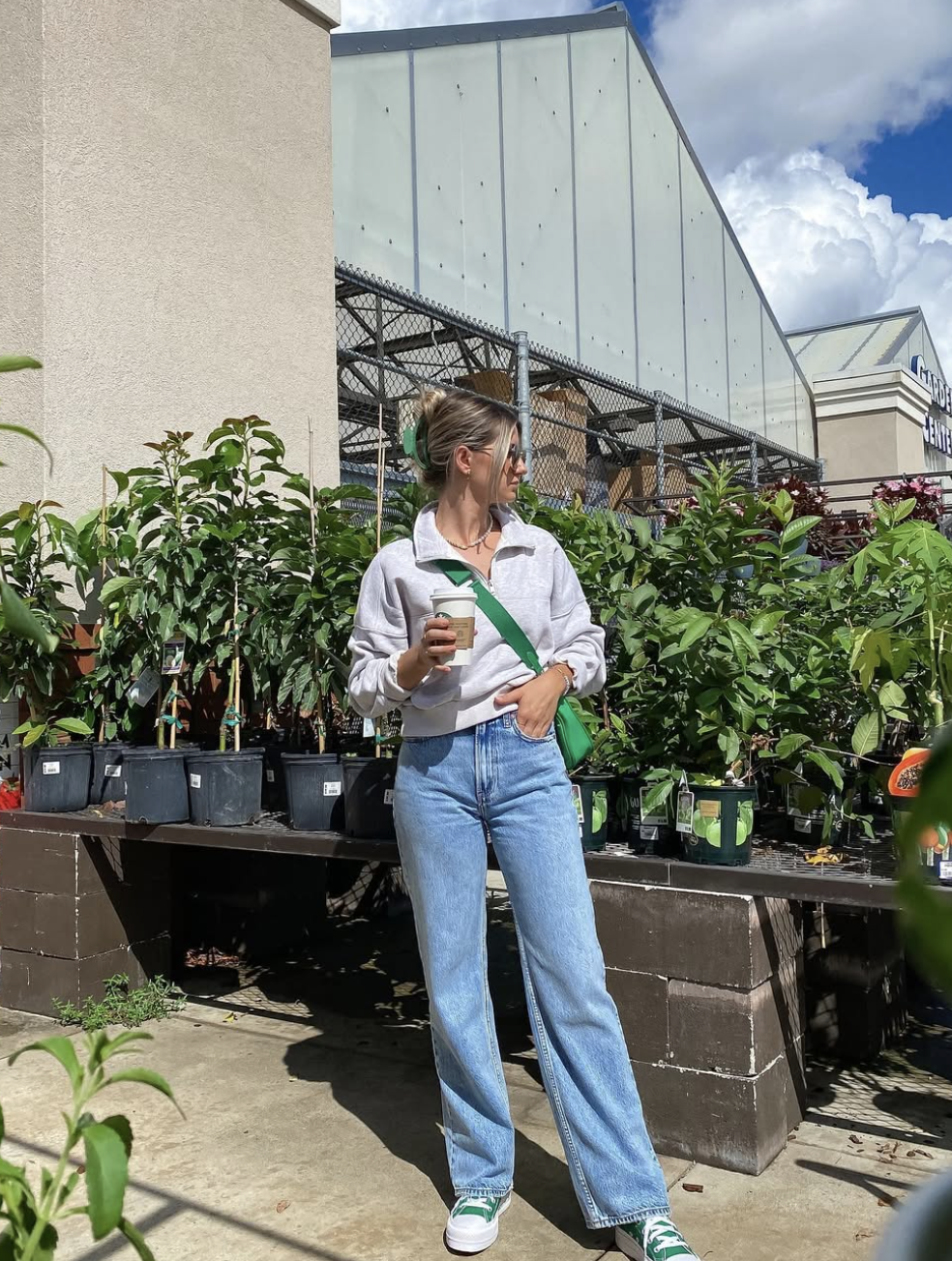 Woman wearing a light grey quarter-zip sweatshirt, high-waisted wide-leg jeans, and green sneakers, carrying a vibrant green crossbody bag at a plant nursery.