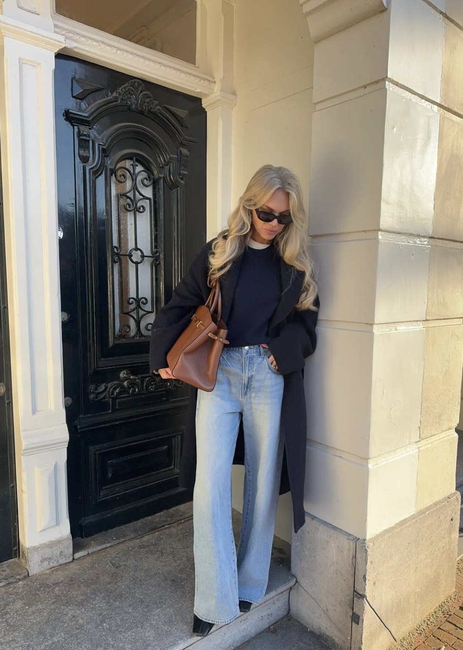 Fashion influencer wearing a long black coat over a navy blue sweater and light wash bootcut jeans with a brown leather tote bag.