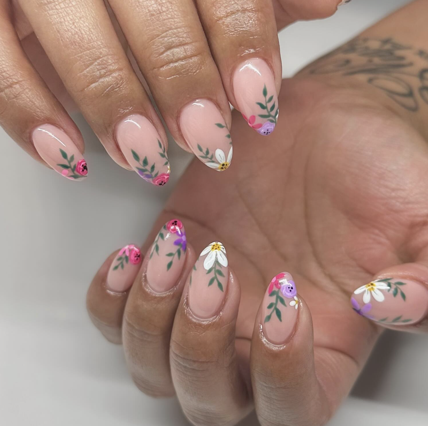 Oval nude nails featuring hand-painted wildflowers with prominent sage green leaves and tiny purple and pink accents.