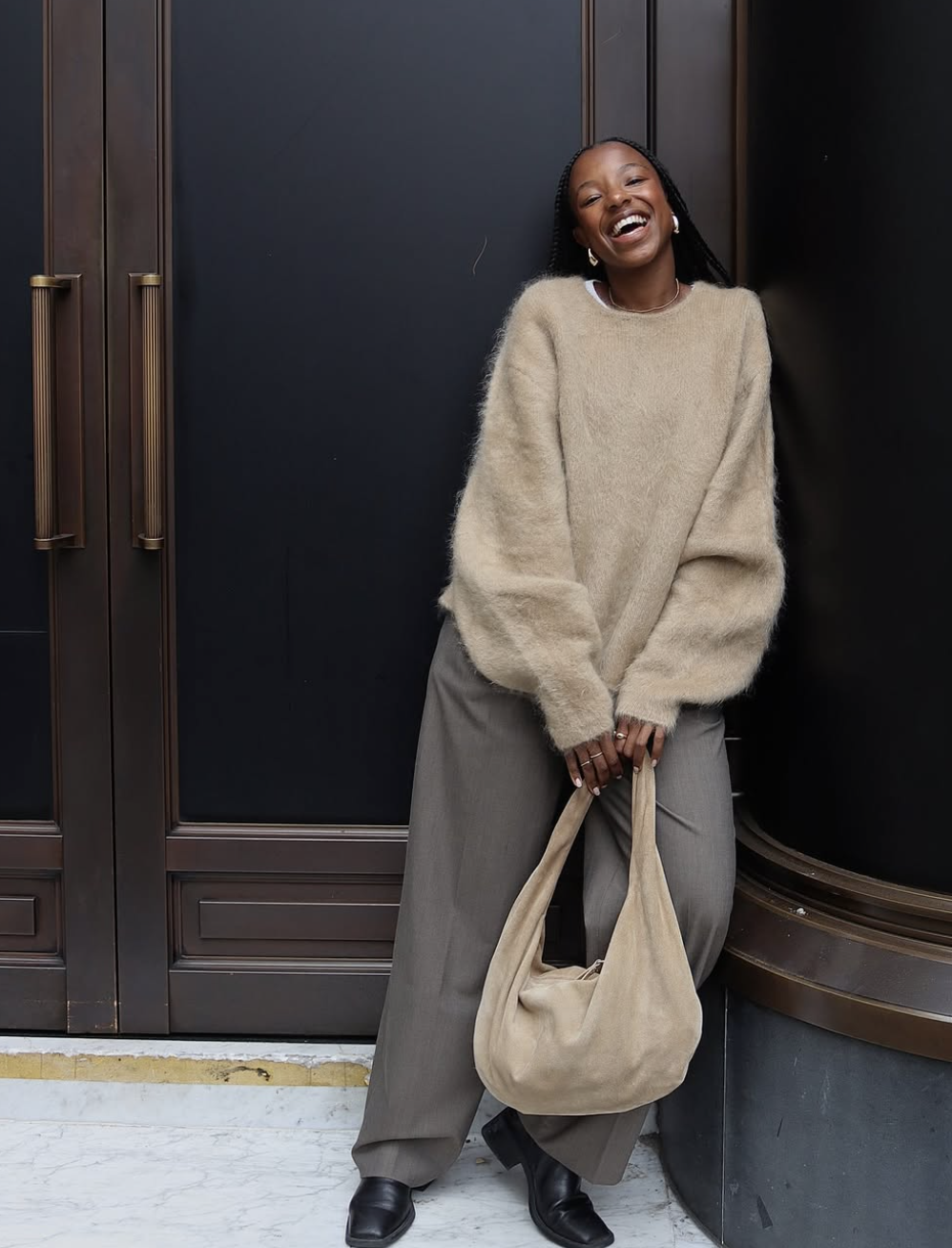 A woman wearing a cozy neutral outfit featuring an oversized chunky oatmeal knit sweater tucked into grey wide-leg tailored trousers, carrying a large beige suede tote bag.