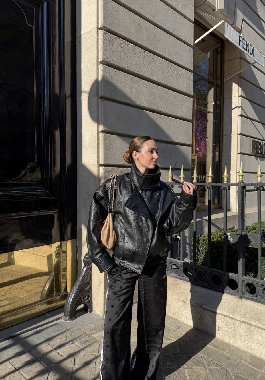 A woman standing on a city sidewalk wearing a structured black leather bomber jacket paired with loose black and white polka dot pants, a tan shoulder bag, and her hair in a sleek bun.
