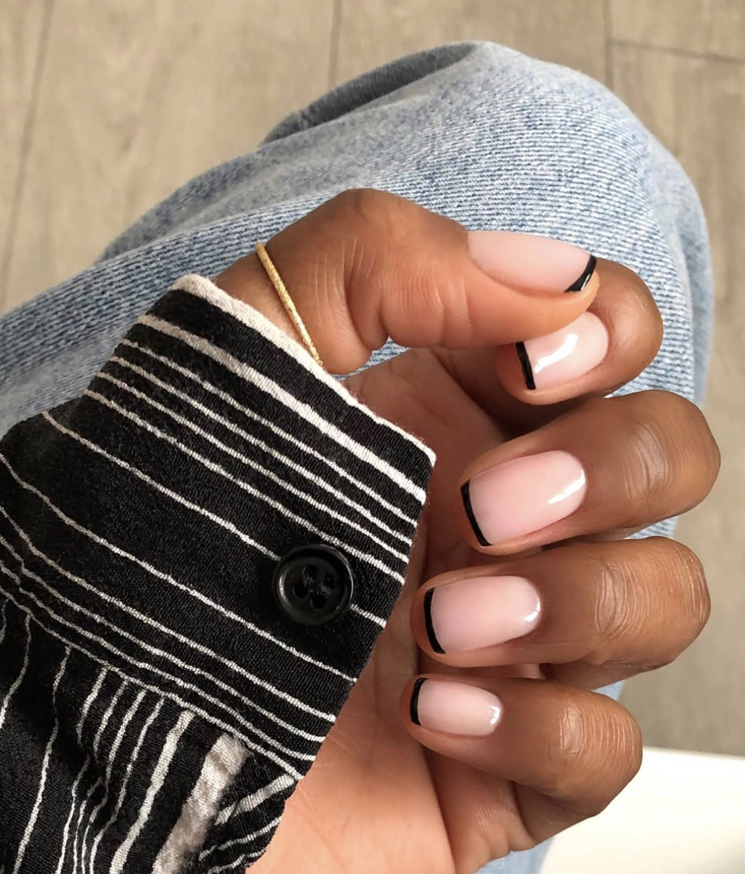 Close-up of a hand with a minimalist micro French manicure featuring ultra-thin black tips on a sheer pink base, styled with a gold ring and a black and white striped sleeve.