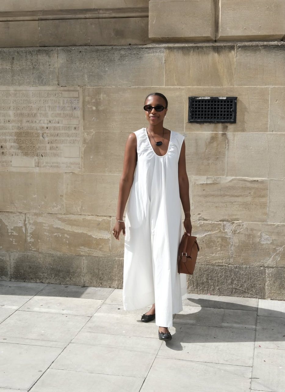 Street-style shot of a woman wearing a voluminous, sleeveless white cocoon maxi dress with a low scoop neckline, styled with a brown leather shoulder bag, black ballet flats, a chunky black pendant necklace, and black sunglasses.