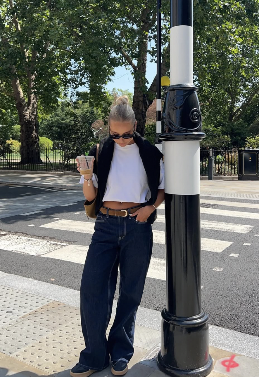 A woman wearing a white boxy crop tee with a black sweater draped over her shoulders, paired with dark wash baggy jeans and a brown belt.