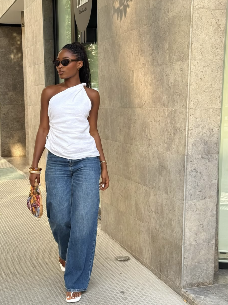 A woman wearing a white asymmetrical one-shoulder tank top with a knotted strap, styled with wide-leg blue jeans and dark sunglasses.