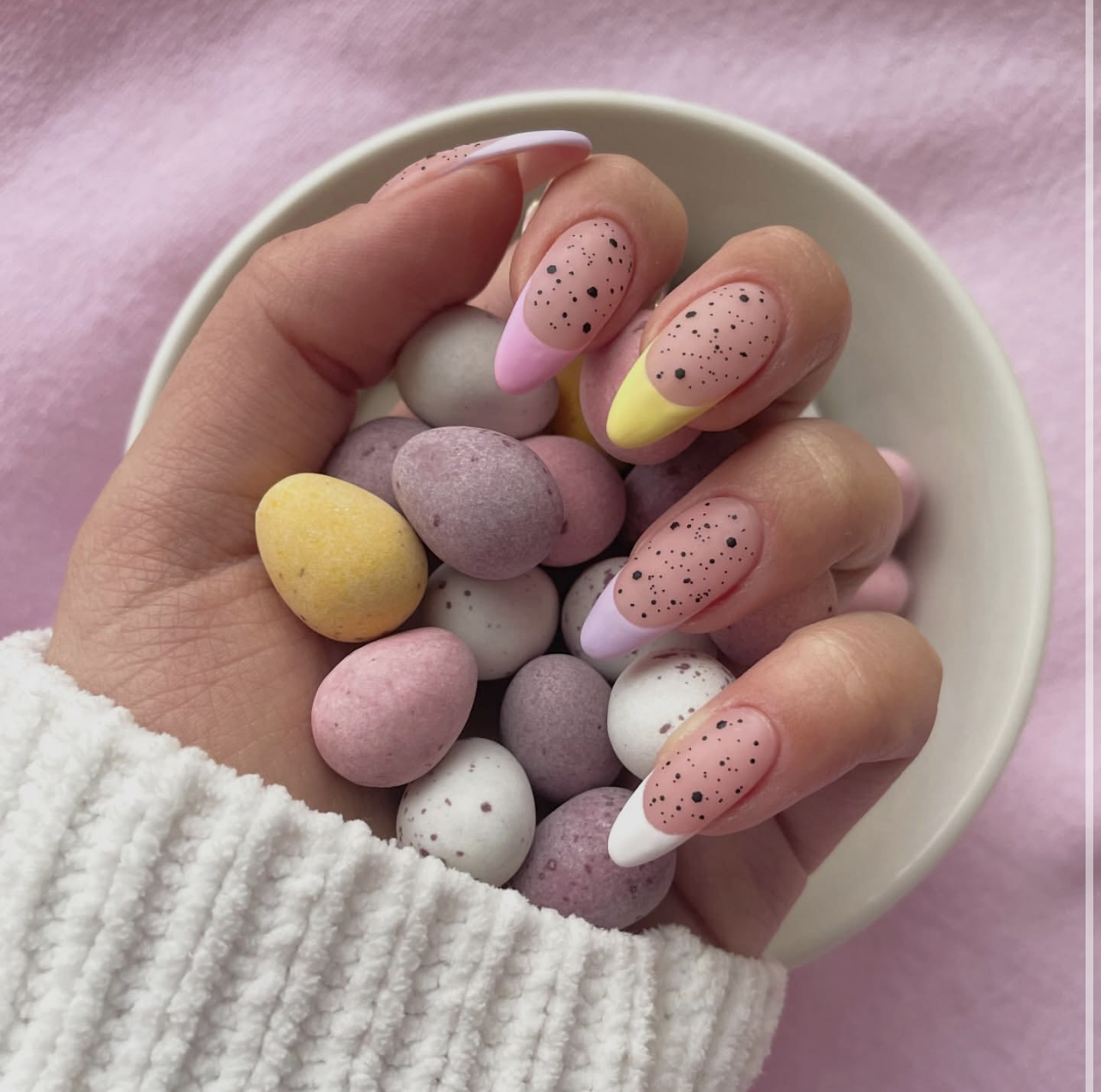 A close-up of a hand with long almond-shaped nails featuring matte-finish pastel French tips in yellow, pink, and purple, covered in black "mini egg" speckles and held against a pile of Cadbury Mini Eggs.