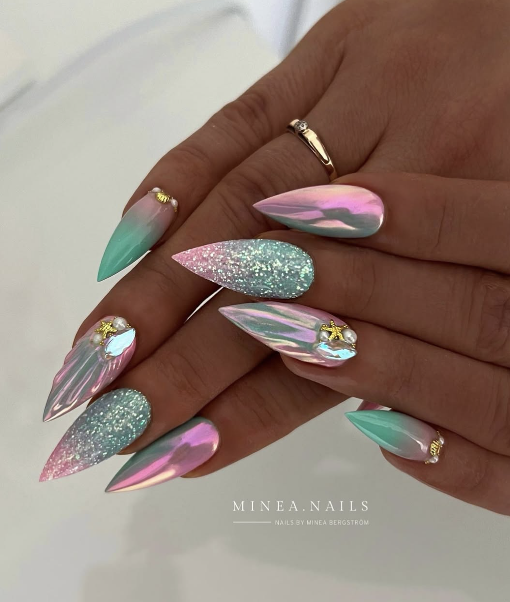 A close-up of long, stiletto-shaped nails with a mesmerizing iridescent pink and mint green chrome gradient, featuring 3D molten silver textures, tiny gold starfish, and small pearl accents.