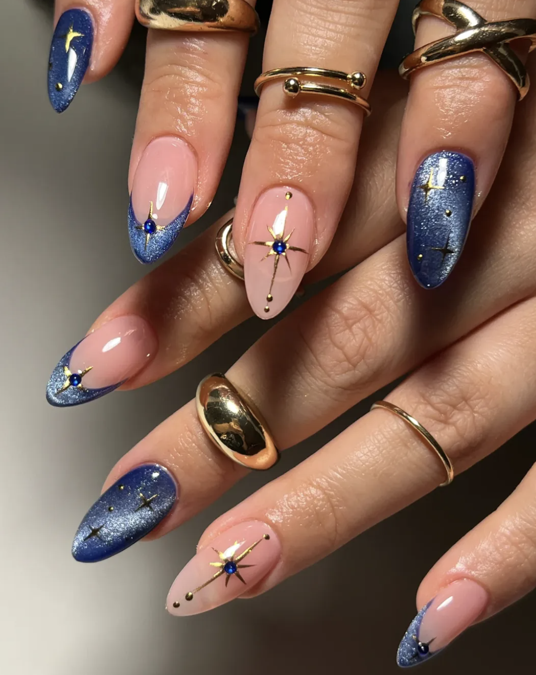 Close-up of extra celestial nails on almond-shaped tips, featuring a mix of shimmering blue cat-eye polish with gold star accents and neutral-toned nails with delicate metallic starbursts.