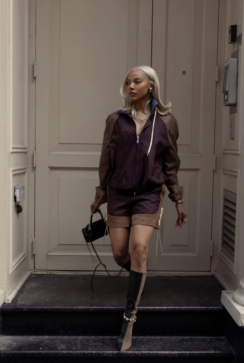A woman with blonde hair wearing a brown and purple zip-up jacket matching shorts set and knee-high heeled boots, holding a small black handbag.