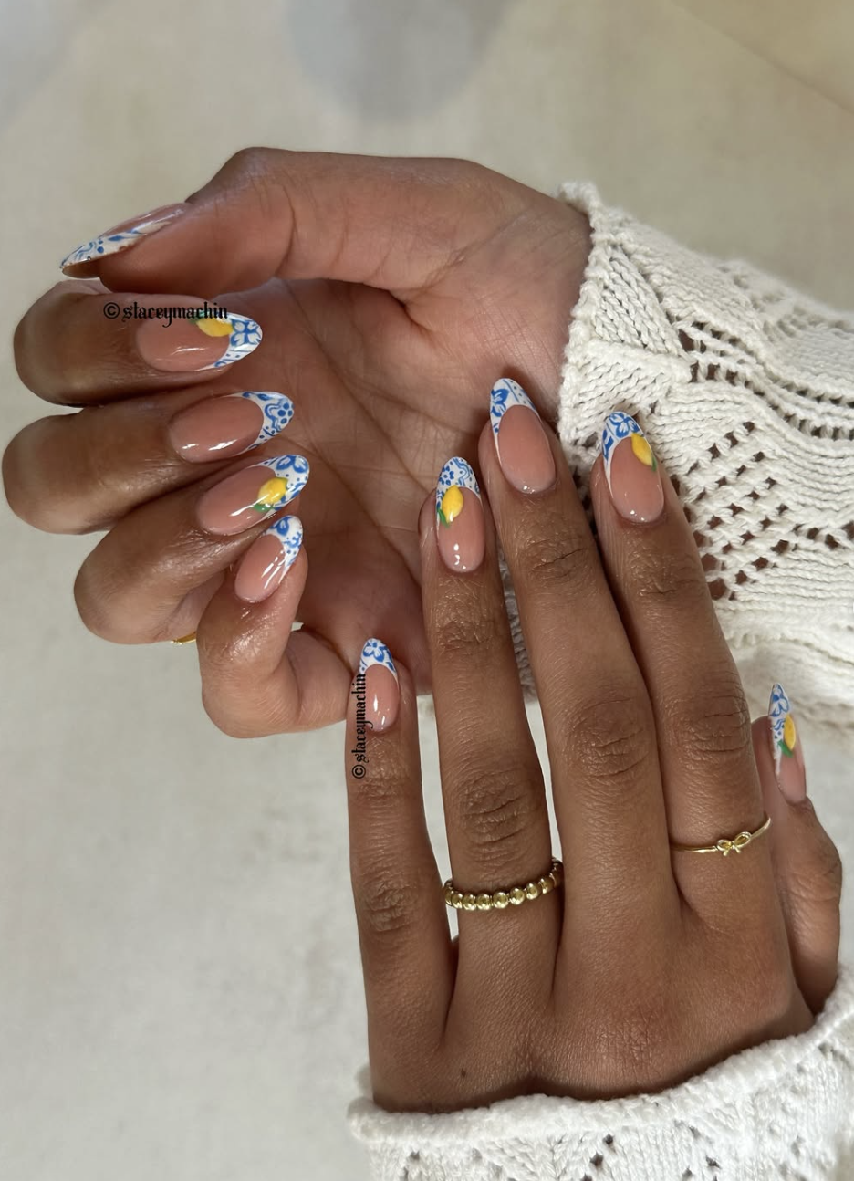 A close-up of almond-shaped nails featuring a minimal nude base with white and blue Amalfi-coast inspired tile patterns and miniature lemon accents hand-painted on the tips.