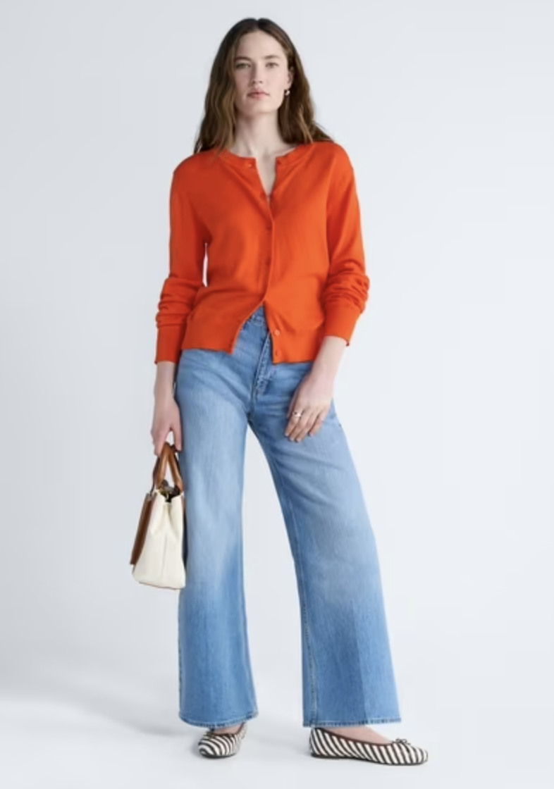 A woman styling a bright orange crewneck button-front cardigan shirt with high-waisted wide-leg light blue denim jeans and holding a cream and brown leather tote bag.