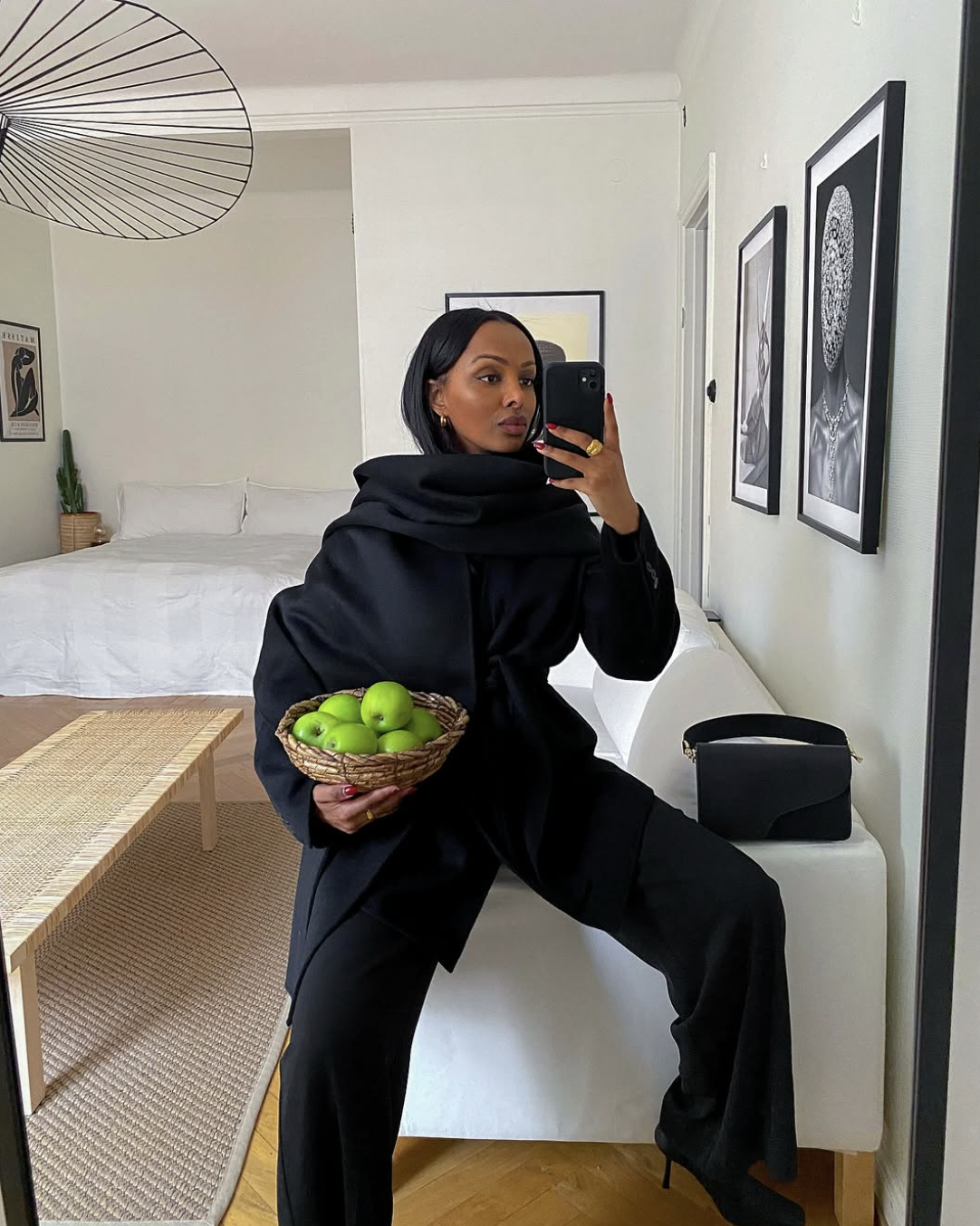 A woman wearing a monochromatic all-black outfit consisting of a structured blazer and trousers, holding a basket of green apples to show contrast.