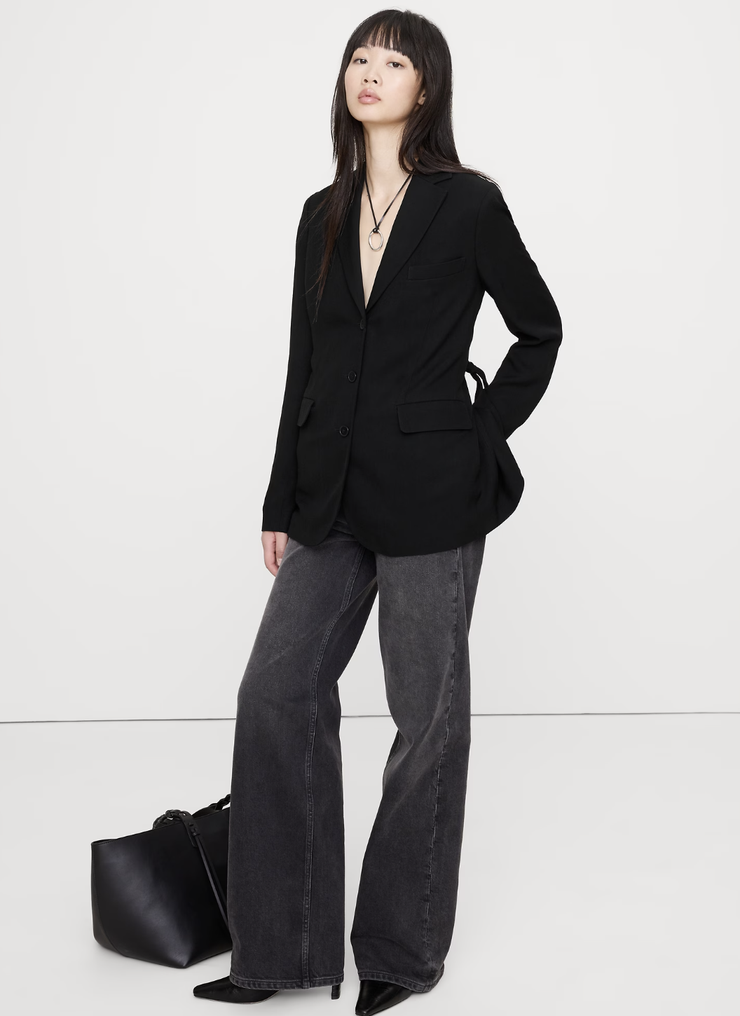 A woman wearing a black tailored blazer with a matching necklace over wide-leg grey denim and pointed black heels.