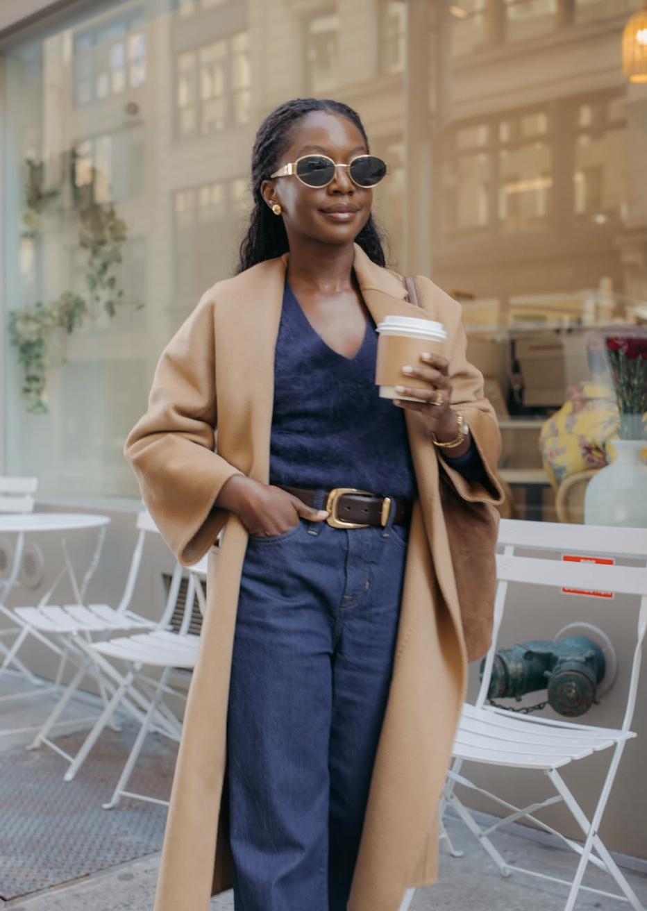 A woman wearing a floor-length camel coat over a navy knit top and wide-leg jeans, holding a coffee cup.
