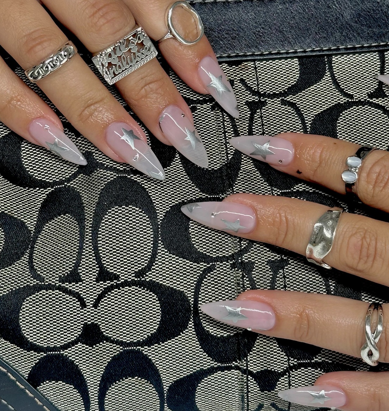 Long pointed almond nails with a sheer nude base featuring silver star decals and small crystal accents.