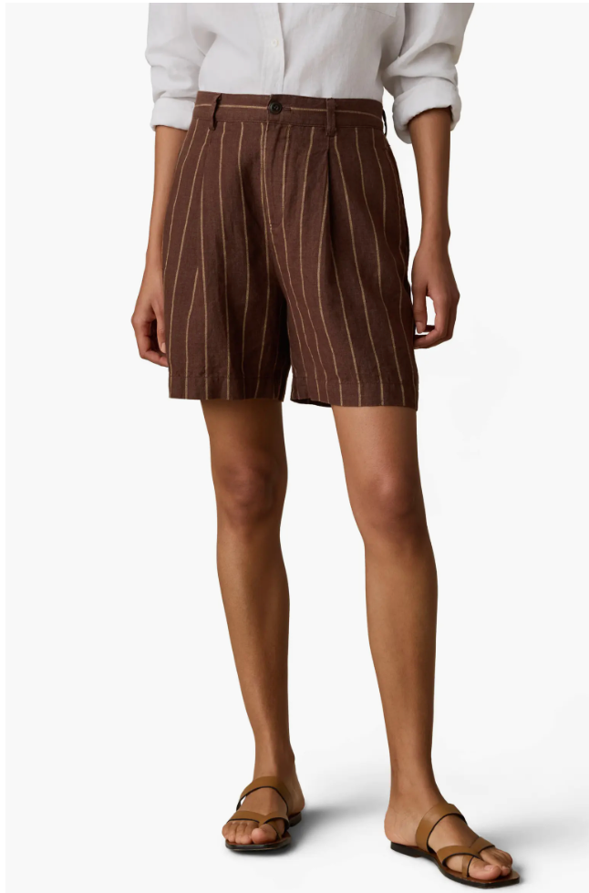 A close-up view of a woman styling high-waisted, structured brown trouser shorts with subtle pinstripes and a relaxed, wide-leg fit, paired with a white button-down shirt.