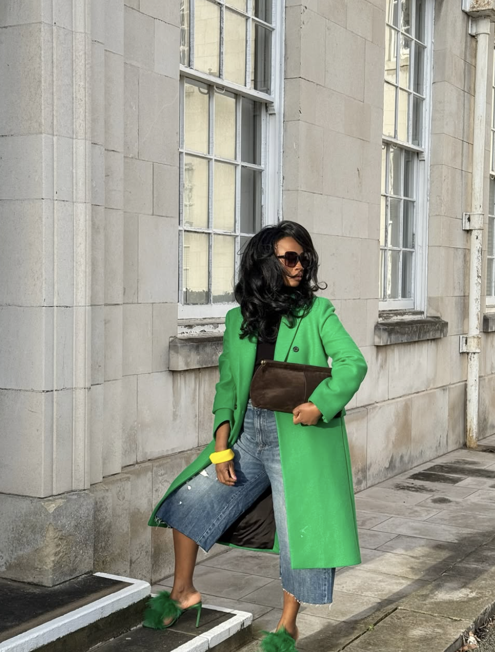 A woman wearing a long bright green coat over a black top and denim Bermuda shorts, paired with green fur-trimmed strappy high-heeled sandals and a brown clutch.