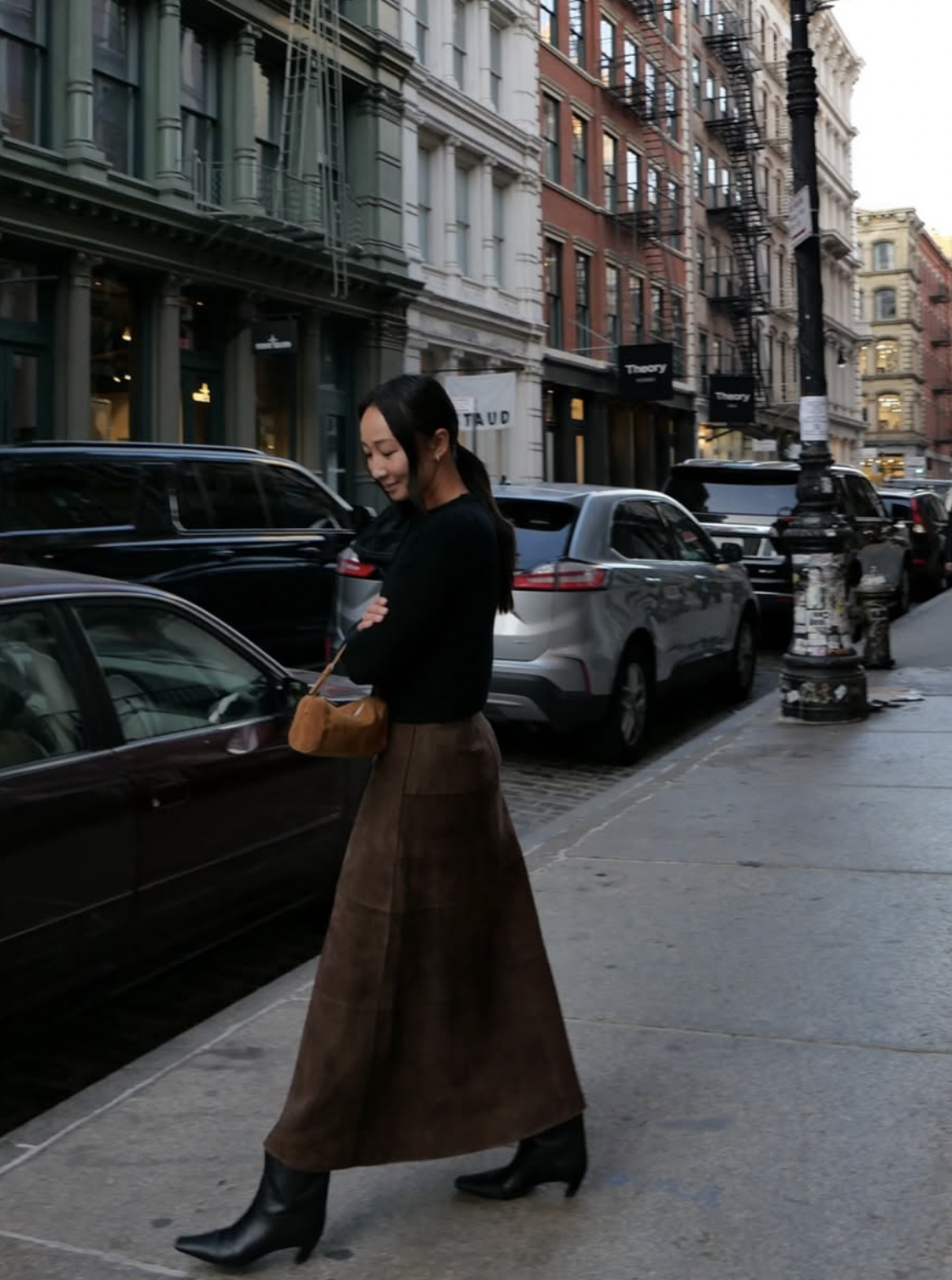 Woman wearing a brown suede midi skirt with a black sweater and black ankle boots on a city street