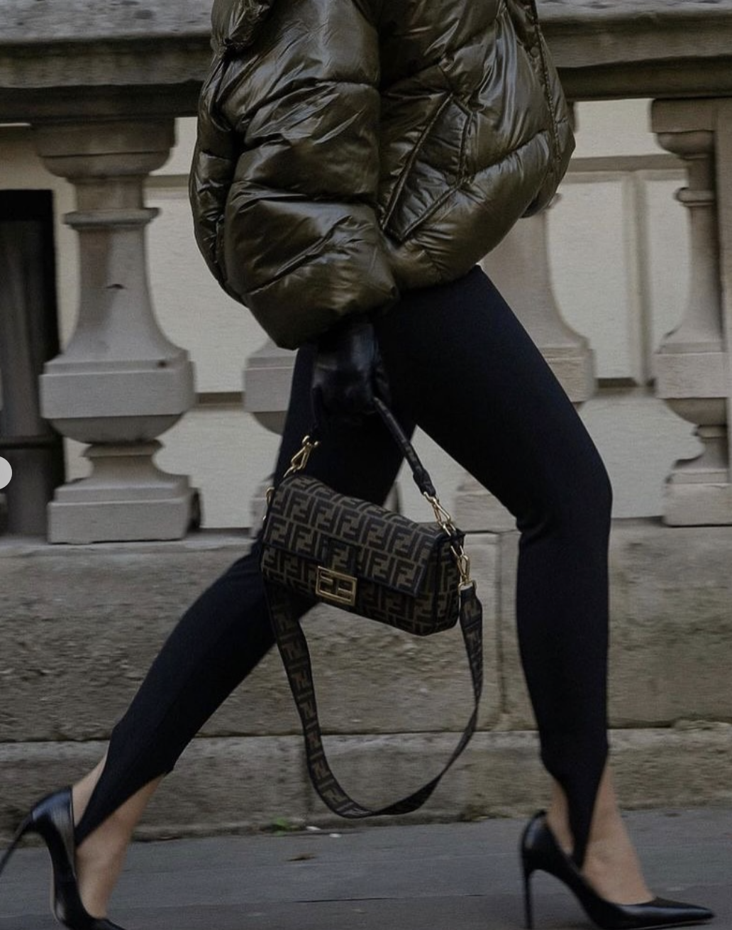 Woman wearing black stirrup leggings with a cropped olive green puffer jacket and black pointed-toe stiletto heels, carrying a patterned Fendi baguette bag.