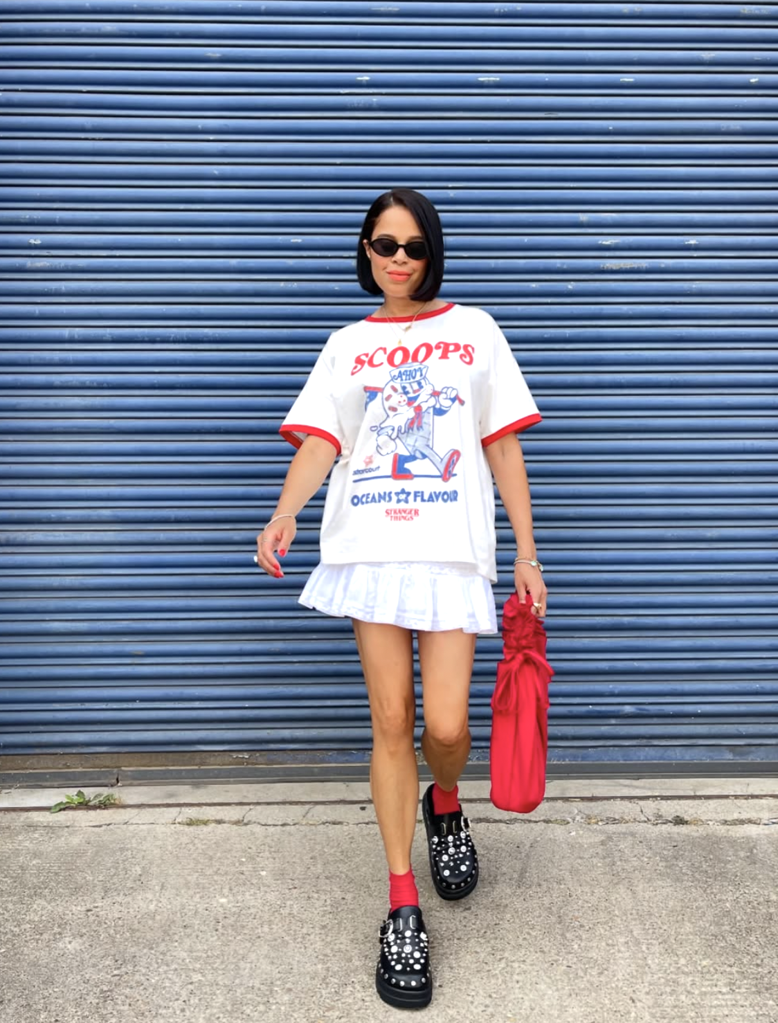Woman wearing an oversized white Scoops graphic t-shirt with a red ringer neck, a white pleated mini skirt, red socks, and black pearl-embellished loafers.